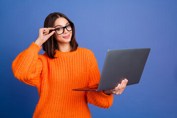 Smiling woman in orange sweater holding laptop while adjusting glasses in front of a purple...