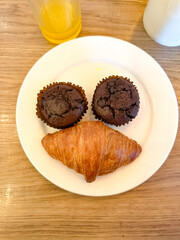 Two chocolate muffins and a croissant arranged on a white plate resemble a smiling face, adding whimsy to a cozy breakfast scene with drinks on the side.