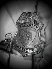 Close-up of a private security officer badge clipped to a white uniform. The badge features an emblem with scales of justice and a lion in the center. Next to the badge is a metallic whistle. 