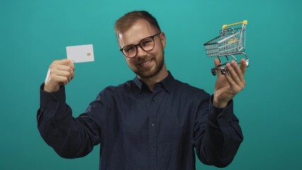 Man smiling and holding a white creditcard in one hand and a miniature shopping cart in the other in studio with turquoise backdrop; confidence savings finance.