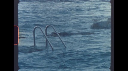 Metal ladder leading into the sea as ocean waves crash over rocks shot on analog 8mm film with a retro nostalgic feel