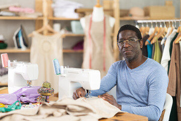 Skilled middle-aged male clothing designer stitching beige linen fabric using sewing machine in creative studio, surrounded by tailoring supplies