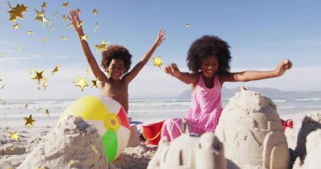 Laughing siblings wearing swimwear raising arms beside sandcastles at beach, with golden confetti