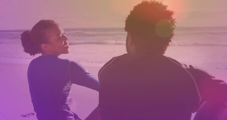 Smiling surfer couple wearing wetsuits sitting at sandy beach during sunset, with lens flare
