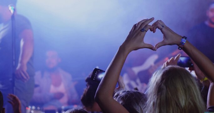 Forming heart shape woman wearing sleeveless top at live music venue, with phones and stage lights - Powered by Adobe