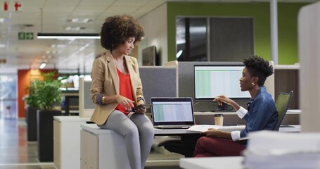 Discussing two colleagues in business casual sharing data on tablet and laptops in open-plan office