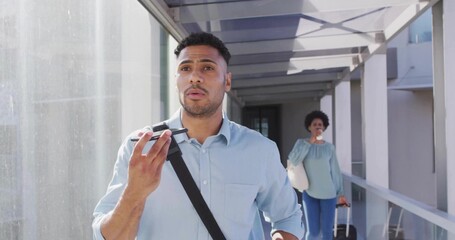 Hispanic man speaking into smartphone in skybridge, with woman drinking coffee and pulling suitcase