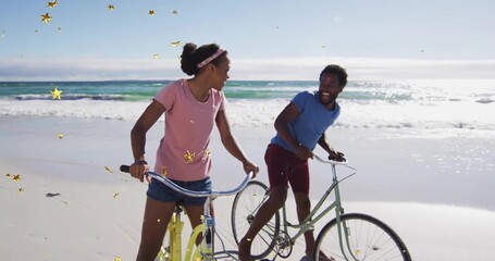 Walking two friends pushing pastel bicycles on sandy beach, smiling amid gold star-shaped elements