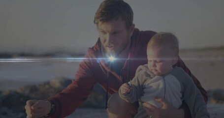 Crouching father holding baby boy pointing toward wet sand on beach in afternoon, with small shell