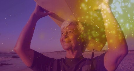 Lifting senior surfer raising surfboard overhead on sandy beach at sunset, with sparkling glow