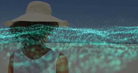 Standing woman wearing straw hat, white top at dusk beach, with glowing turquoise particle overlay