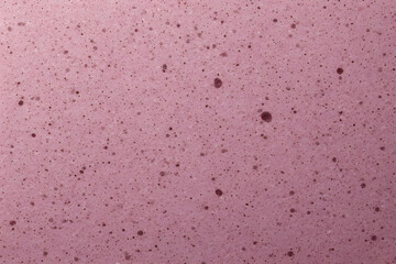 Vibrant pink or rosy abstract background with a uniform, porous texture, densely covered with tiny, dark speckles and larger dark spots, resembling berry-flavored foam.