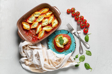 Baking dish and plate of stuffed conchiglie pasta with cheese, spinach, tomato sauce on grey background
