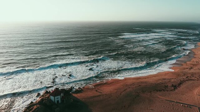 Wide drone shot showing Capela do Senhor da Pedra chapel by the ocean waves at Miramar beach near Porto Portugal