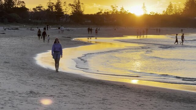 model girl in elegant shirt walks in shallow water at beach admiring stunning orange sunset behind mountain ranges at popular Byron Bay Beach, New South Wales, Australia