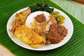 Delicious Padang rice dish with spicy beef rendang, omelette, cassava leaves, and sambal green chili, served on banana leaf background.