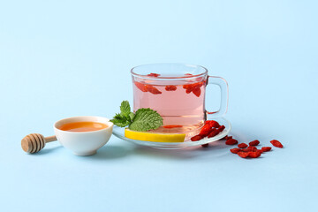 Glass cup of hot goji tea with mint and bowl of honey on blue background