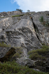 Steep granite wall with cracks and vegetation at Bowman Valley, California, USA. The rock formation rises vertically against a partly cloudy sky, featuring rugged textures and ledges popular among roc
