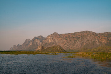 The mountain looms over a green landscape where a swamp meets a tranquil lake, water reflecting the sky, a single cloud drifting in peaceful, untouched nature.