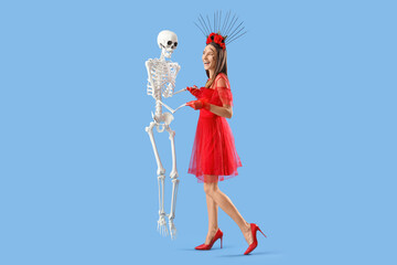 Beautiful young happy woman dressed for Halloween dancing with skeleton on blue background