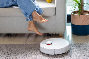 Young adult woman lifting feet while robot vacuum cleans carpet in living room