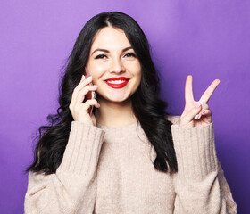 Beautiful smiling brunette woman talking on mobile phone and showing v-sign over purple background.