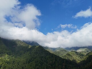 Aerial view of a lush green mountain covered with dense tropical forest under a bright blue sky and low clouds.