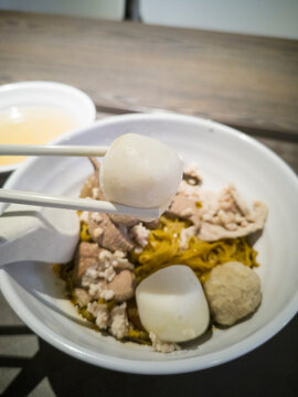 One big round handmade fishball is picked up by a pair of chopsticks in the foreground against blurred background of a bowl of minced meat noodle in an eatery