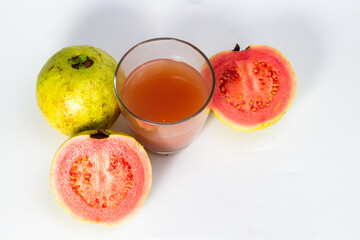 A glass of fresh guava juice is placed among whole and halved guavas on a white surface.