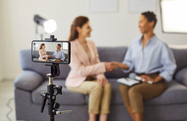 Two women sit on a sofa and shake hands while filming a brief interview, the recording made on a...