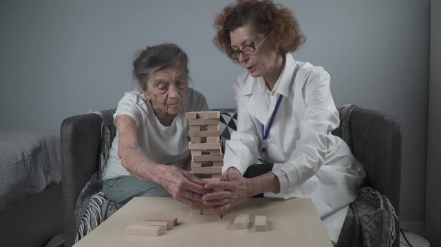 Jenga game. Theme is dementia, aging and games for old people. Caucasian senior woman builds tower of wooden blocks with the help of a doctor as part of a therapy and jenga game at a patient's home.
