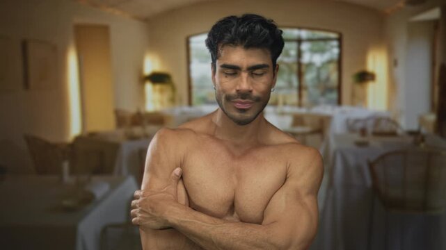 Toned young man hugging bare chest with closed eyes in restaurant interior; comfort calm solace healing.