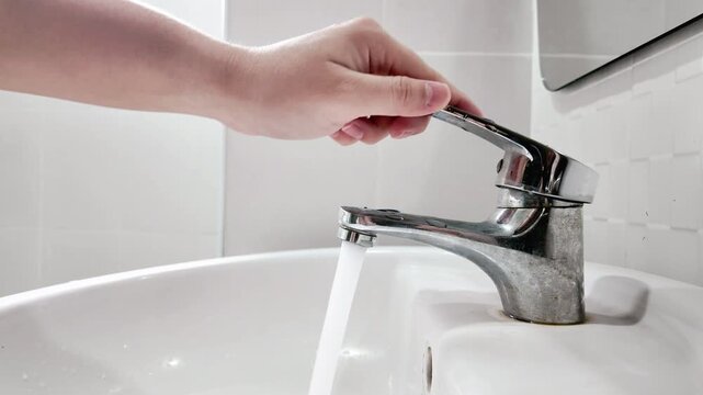 Close-up view of hand turning off silver metal faucet that was left open in clean white ceramic sink in bathroom. Water saving concept.