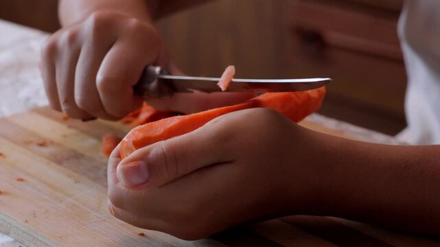 Young beginner demonstrating precise slicing of vegetable with small knife, Child at home practicing careful carrot peeling and cutting using tiny kitchen knife