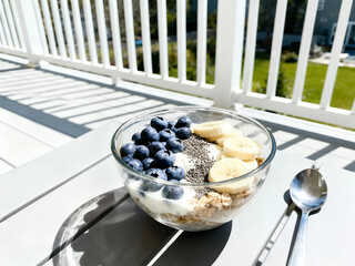 A healthy breakfast bowl with Greek yogurt, granola, fresh blueberries, and banana slices is topped with chia seeds on a sunny outdoor patio table.