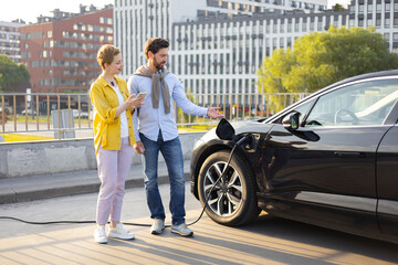 A couple stands near their electric vehicle while it charges at a charging station in an urban setting.