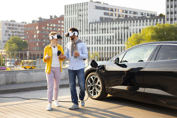 A couple wearing VR headsets walks near a charging electric vehicle in an urban setting.