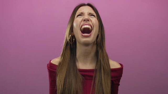 Woman with brown hair wearing hoop earrings opens mouth wide and tilts head back in pink studio; anger.