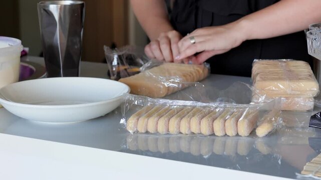 Caucasian woman opening packs of ladyfinger biscuits on counter, rows of cookies and empty packaging beside white bowl, methodical prep for layered dessert, bright
