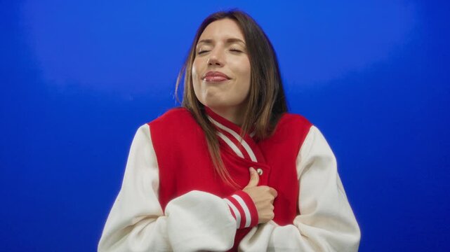 Woman cheerleader in varsity uniform grips red jacket collar in vibrant blue studio while smiling gently with eyes closed and relaxed posture; confidence pride.