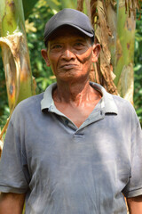 A seasoned farmer stands among tall banana trees, gazing directly at the camera with a calm and steady expression under natural sunlight, representing rural dedication and hard work.