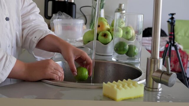 Fresh fruit washing in kitchen, Fruits being cleaned with water and sponge in kitchen, Person rinsing green apple under running water with cleaning tools in bright