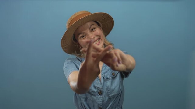 Woman wearing straw hat and denim shirt forming hashtag gesture with fingers in studio against blue background; playfulness.