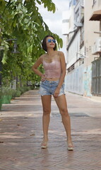 Confident woman in lace camisole and denim shorts poses on sunlit city sidewalk, showcasing bold summer fashion with wedge sandals and reflective sunglasses