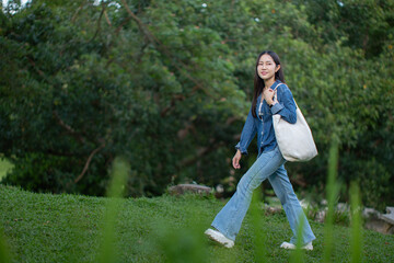 Fototapeta premium A young Asian woman wearing a denim jacket and jeans walks in the park carrying a white canvas tote bag, living an eco-friendly and relaxed lifestyle.