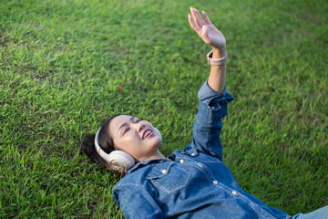 A beautiful Asian woman in a jeans shirt is sleeping lying on the grass, wearing headphones, and raising her hands to the sky happily.