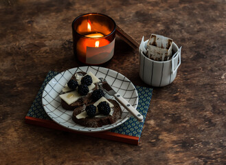 Coffee, toast with cheese, blackberries and honey, a lighted candle, a book on a wooden table. Cozy mood