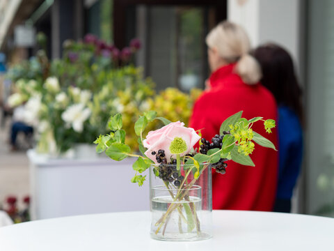 Two customers stand by a flower shop counter looking at fresh orchids lilies and allium arranged in glass vases near an open door. 