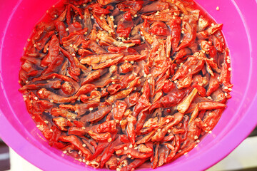 A top-down, close-up view of dried red chili peppers soaking and rehydrating in water inside a vibrant pink plastic bowl, with seeds floating on the surface.