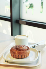 A cinnamon roll and a hot latte on a tray, set against a large, bright window in a modern cafe. A peaceful and minimalist scene for a morning coffee break.
